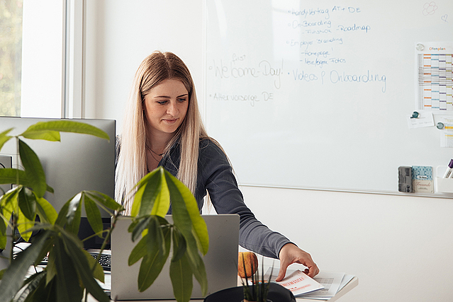 Mitarbeiterin steht an ihrem Schreibtisch und arbeitet am Computer, im Hintergrund ein Whiteboard mit Notizen.