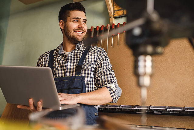 Glücklicher Handwerker mit Laptop in der Werkstatt. Er profitiert vom Empfehlungsmarketing von FirmenABC.